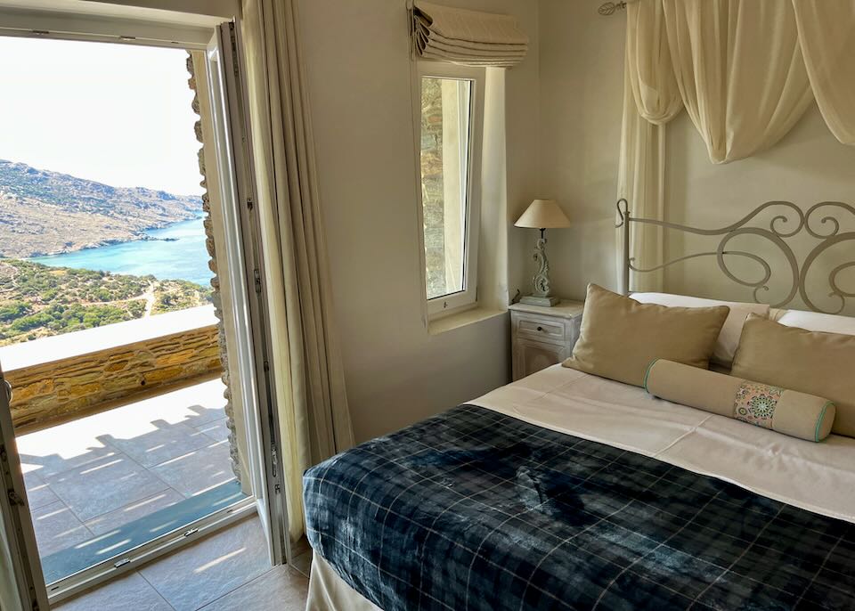 A hotel bed near an open balcony door with view of the beach and mountainous hillside