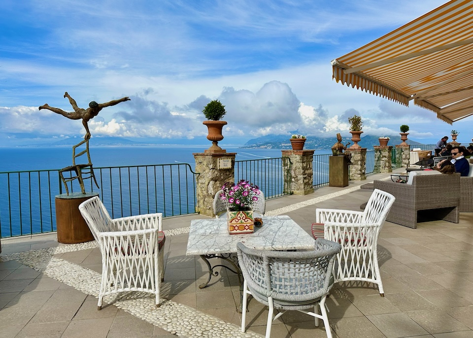 Open-air terrace overlooking the sea from high above with a whimsical sculpture and dining tables under a striped awning at Hotel Caesar Augustus in Anacapri, Capri