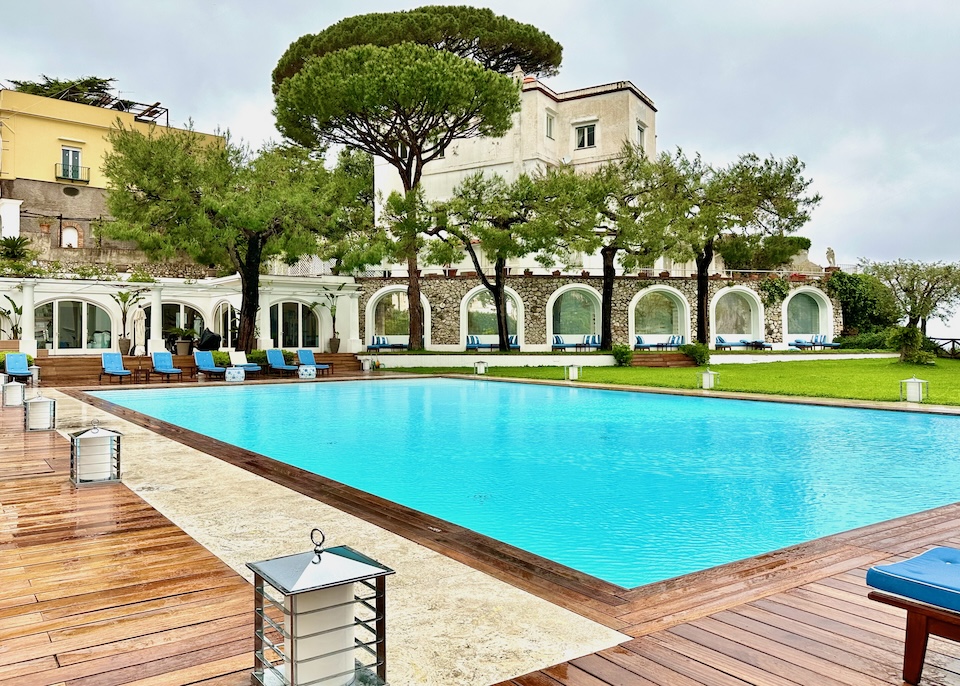 Pool with a wooden deck in front of a building with arched windows and shade trees at JK Place in Marina Grande, Capri