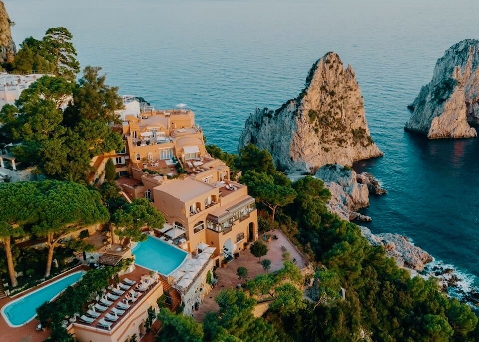 View of apricot-colored Punta Tragera hotel from above looking down on two pools and two dramatic rock formations in the sea in front of the hotel