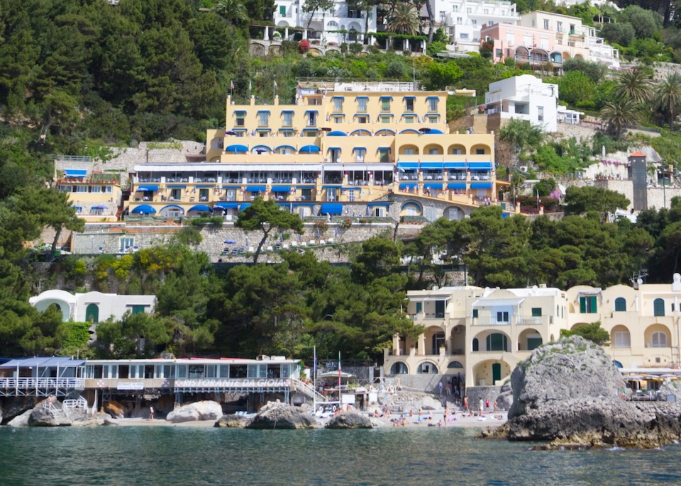 The yellow exterior of Hotel Weber Ambassador with blue awnings on the hill just above the beach at Marina Piccola in Capri