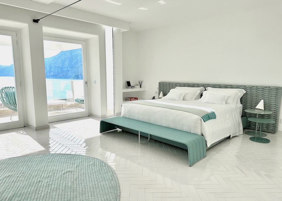 The bedroom of a suite in white and pale turquoise with floor-to-ceiling windows facing the sea at Casa Angelina in Praiano on the Amalfi Coast