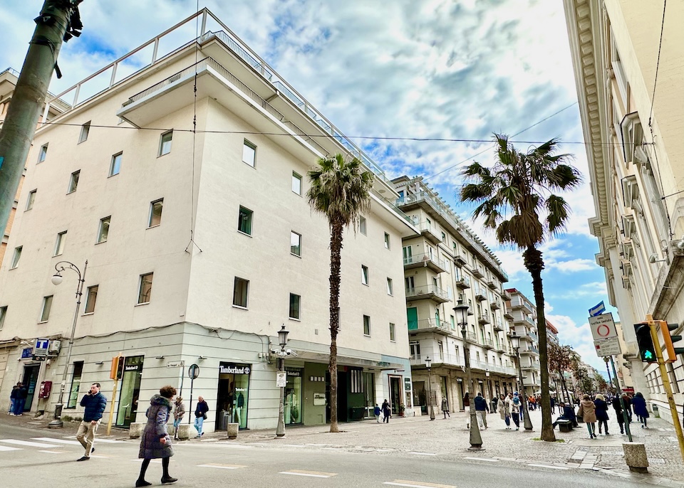 Outside the modern Hotel Montestella with two palm trees flanking a busy walking street in Centro Nuovo in Salerno