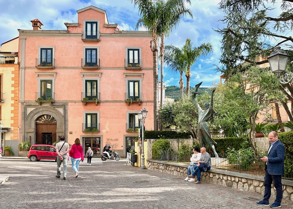 Pink stucco exterior of the Palazzo Marziale with a grand archway entrance and windows with wrought iron French balconies on a cobblestone street near a garden in Sorrento, Italy