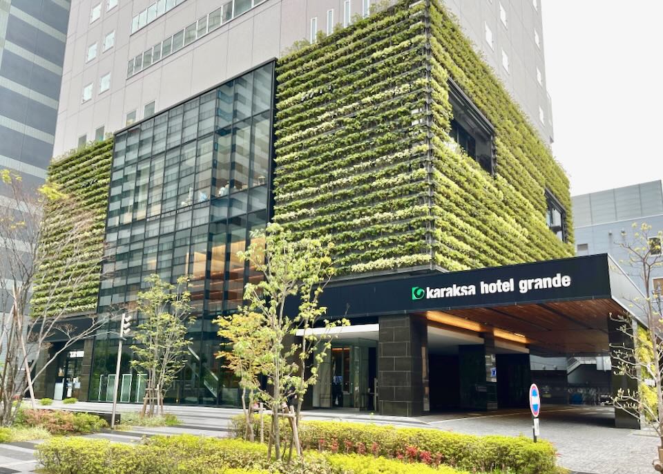 The street entrance to a tall building with plants growing on the first few levels.