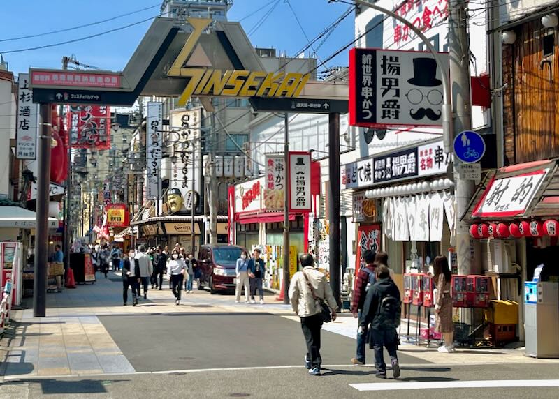 People walk under a sign that says, "Shinsekai," in a busy city area.