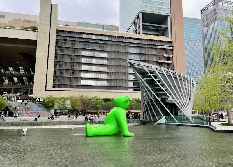 A giant green teddy bear sculpture sits in the middle of a fountain.