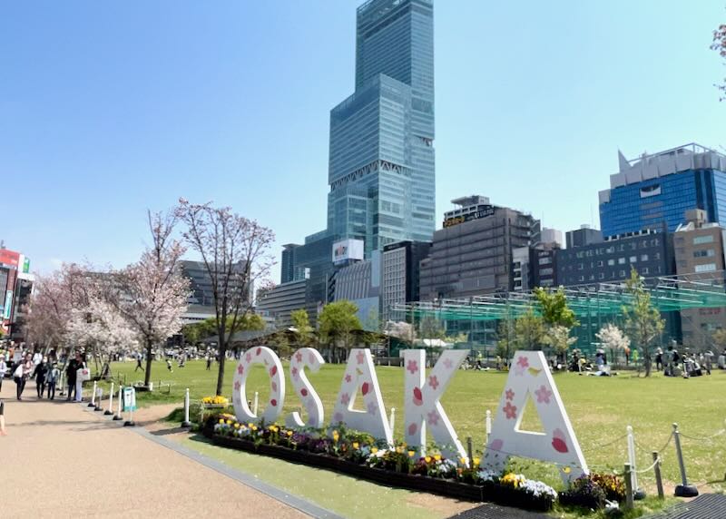 A large sign that reads, "Osaka" sits on the outskirts of a grassy park surrounded by tall buildings.