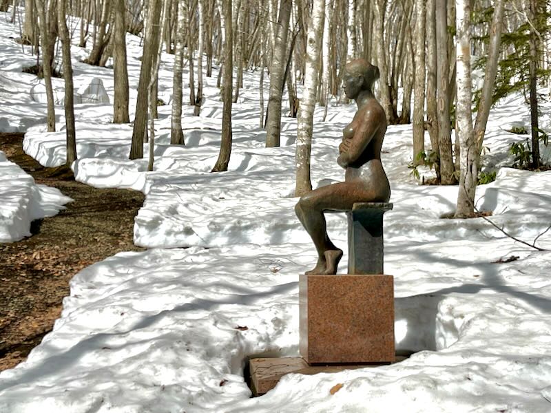 A sculpture of a nude woman sitting on a stool is surrounded by trees and snow.