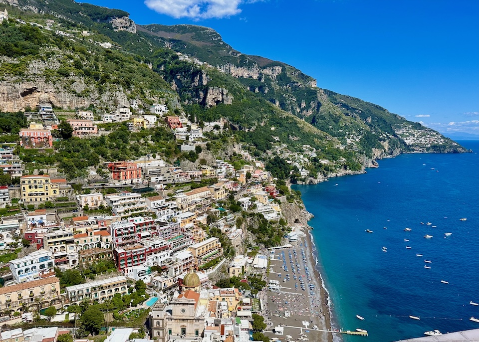 View from the hill toward the sea, beach, church, and town center of Positano with green mountains rising behind.