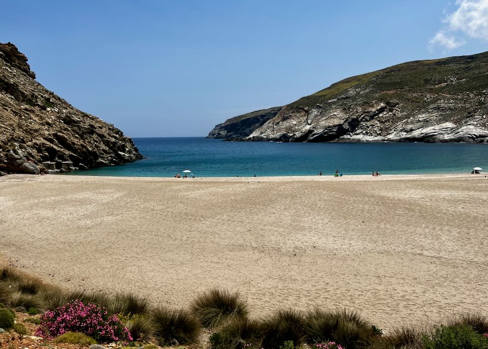Wide sandy beach with blue water and golden sand, tuckedbetween two rocky hills. 