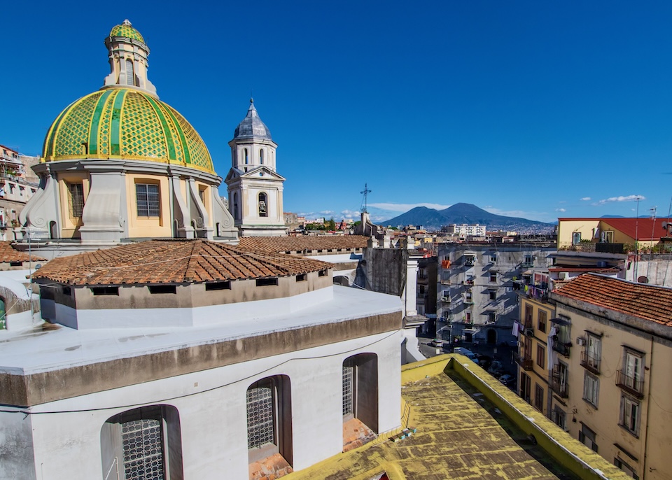 The green and yellow church dome of Church of Santa Maria della Sanità surrounded by red tile rooftops with Mount Vesuvius in the background