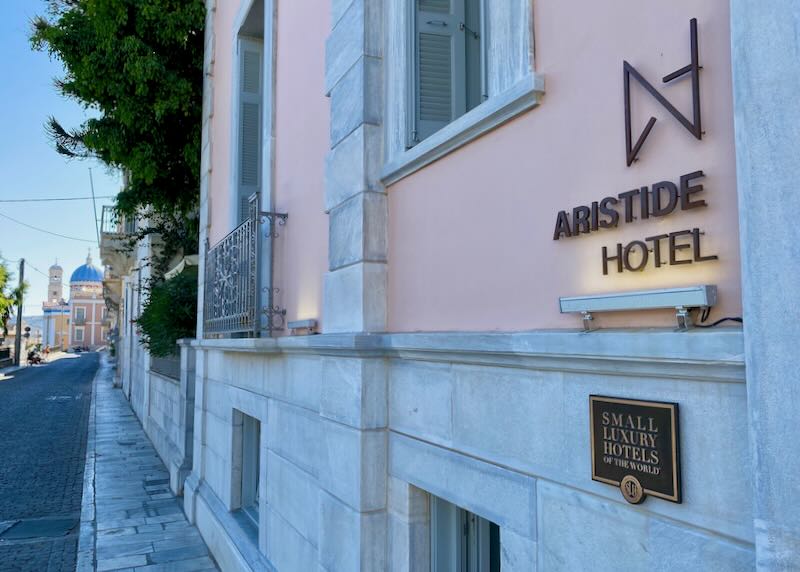 Subtle signage on a neoclassical pink hotel, with a Greek church in the background