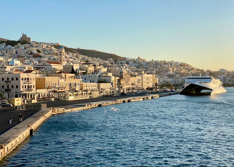 A Greek port town bathed in dawn light, with a hyrdrofoil ferry docked at the waterfront