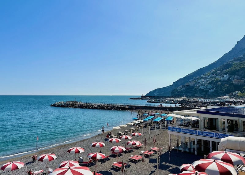 A sandy beach with a pier and red and white striped beach umbrellas in the town of Amalfi