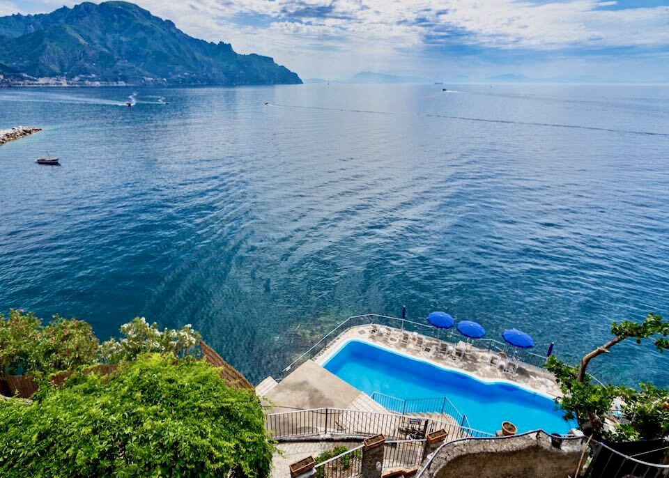 A pool fronted by umbrellas sits at the bottom of a cliff right above the sea at Hotel Luna Convento in Amalfi