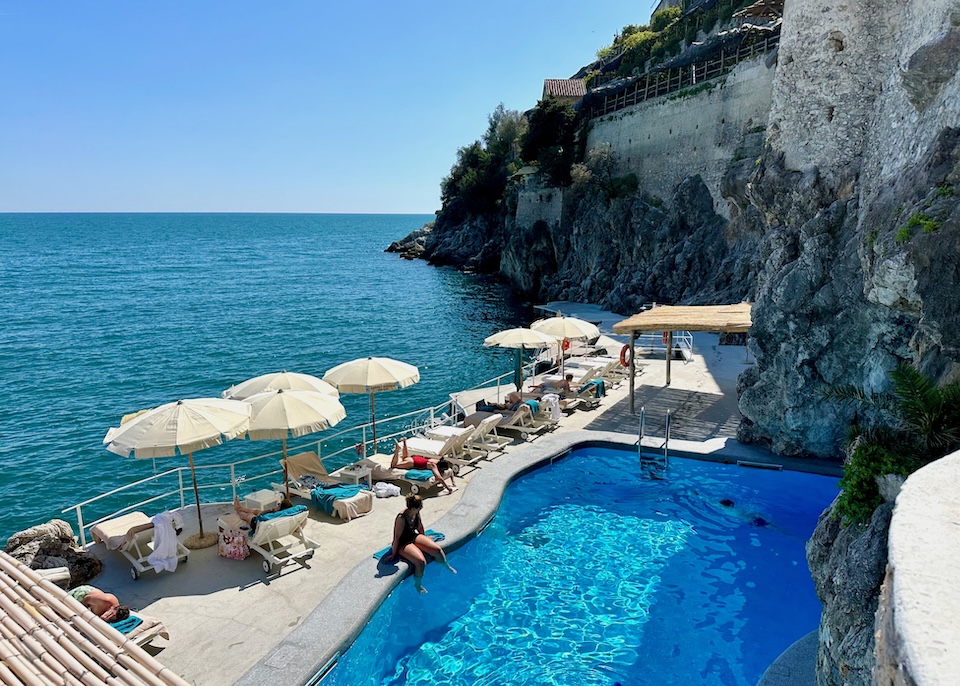 A pool on a seafront terrace with sunbeds and umbrellas at the base of a cliff at Hotel Santa Caterina in Amalfi