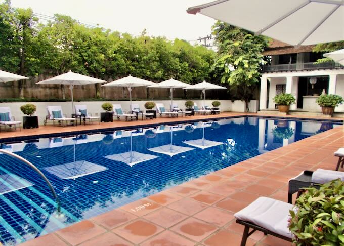 White lounge chairs with white umbrellas sit next to a long blue pool with terracotta tiles.
