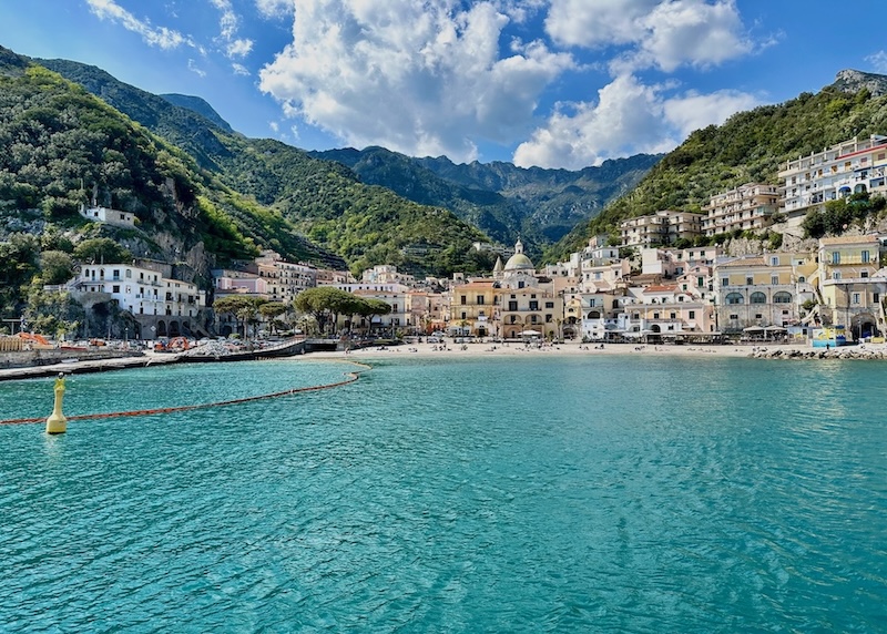 The seaside town of Cetara on the Amalfi Coast as seen from the sea with its beach, domed church, and pastel houses with mountains rising from behind.