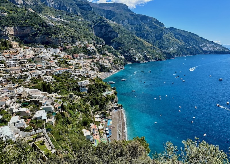Overlooking Positano with two beaches, lush greenery, dramatic mountains, and the sea dotted with little boats.