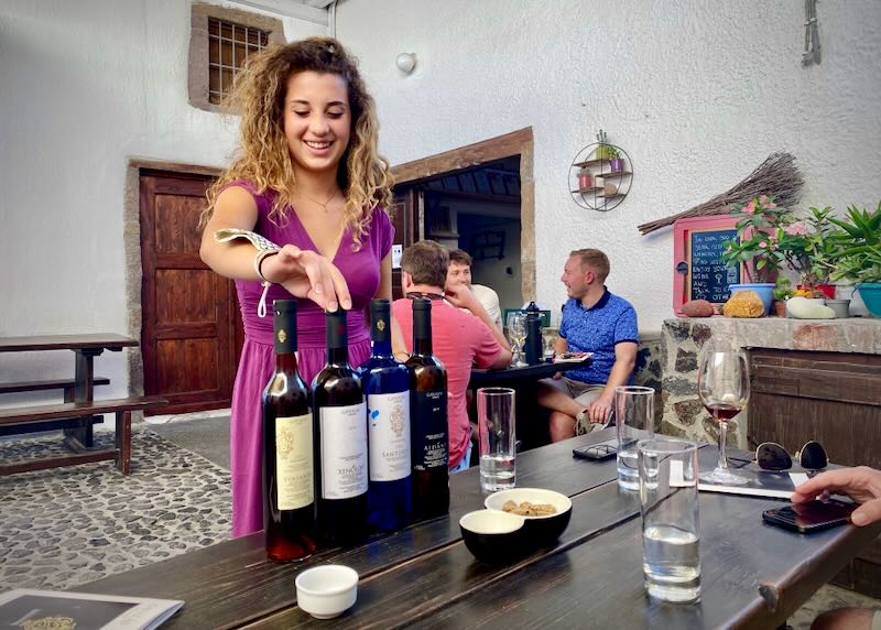 A woman gestures to wine bottles at a wine tasting on a rustic patio
