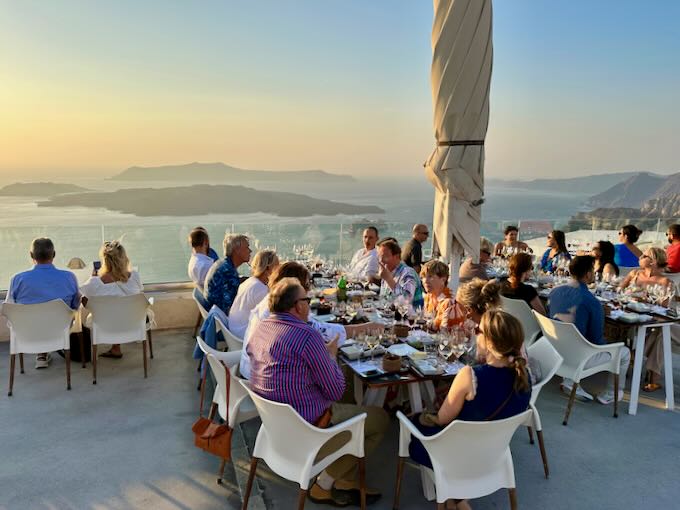 People dine and taste flights of wine on a terrace overlooking a caldera at sunset