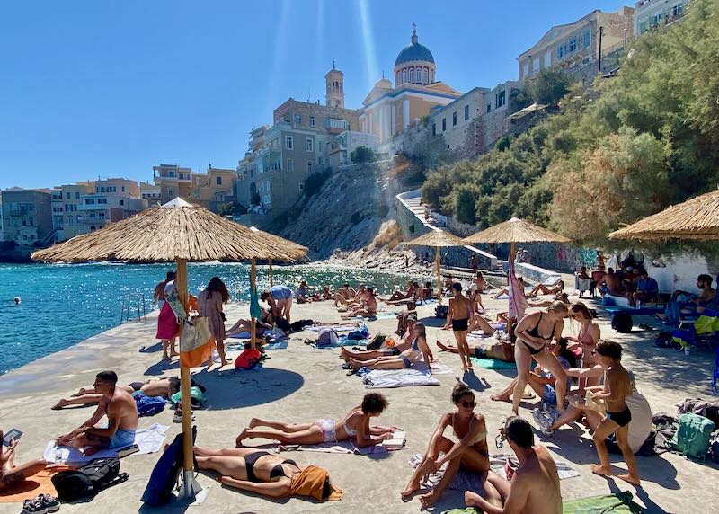 Sunbathers lie under thatched umbrellas on a concrete platform over the sea, with pastel neoclassical buildings in the background