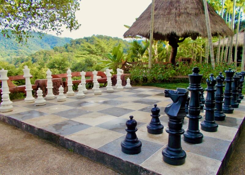 Trees and a thatched hut surround a giant chess board.
