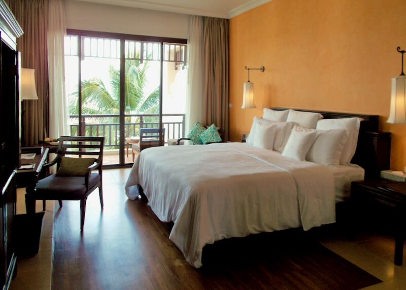 A large white linen bed sits in a hotel room with warm painted walls and natural light from the sidling doors.
