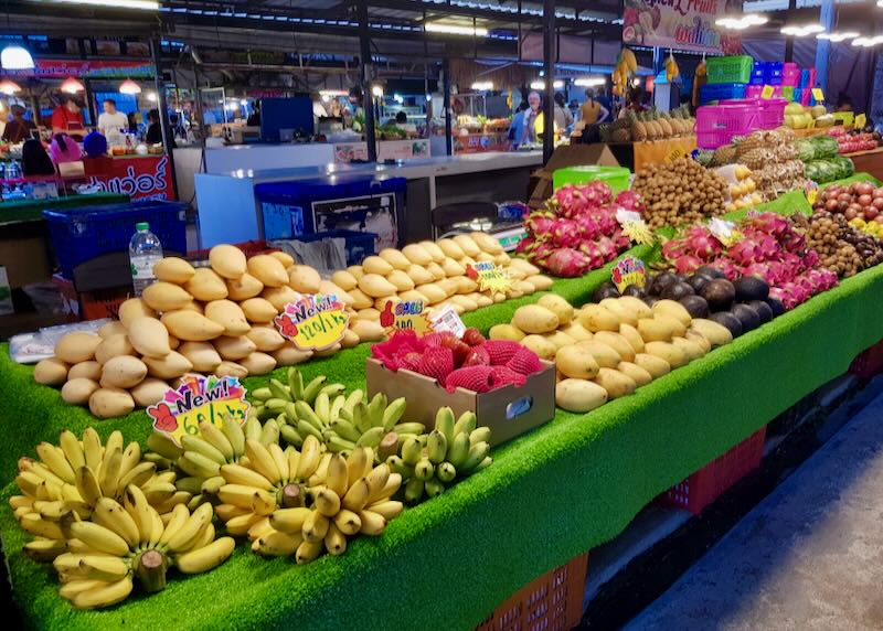 Rows of bright red dragon fruit, yellow bananas, and mangos sit on a table at a market.
