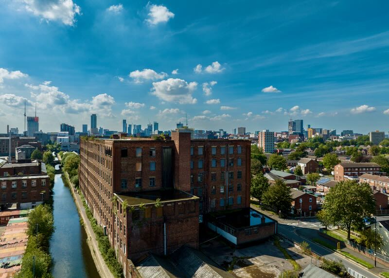 Victorian Cotton Mill in Ancoats, Manchester, England. The frequently used Ashton Canal can be seen running alongside the mill into the city centre. Manchester's modern skyline can be seen in the background. The photograph was produced on a bright sunny day.
