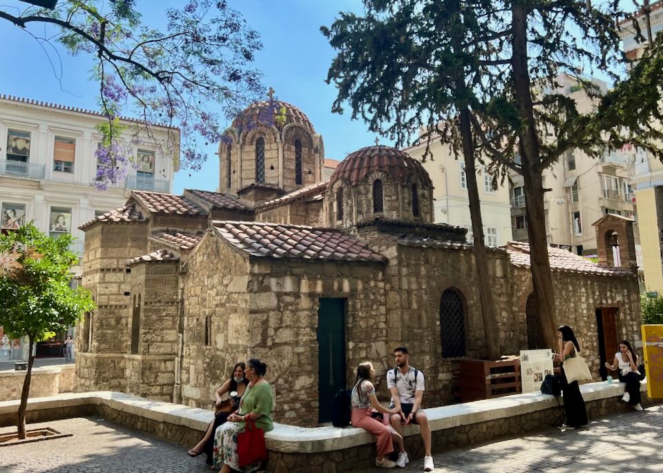 Anterior view of Panagia Kapnikarea church in Athens on a spring day, with people sitting on a low surrounding stone wall.
