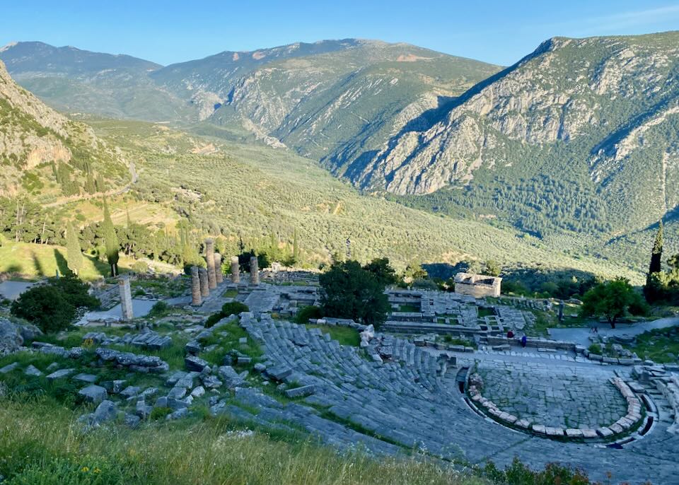 View looking down over ancient Delphi toward olive groves and the mountains beyond, on a sunny day.