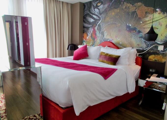 A large black and gold flower mural sits behind a white bed with hot pink velvet pillows and throw blanket.