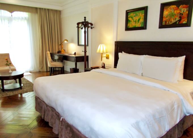 A large bed covered in white linens sits on a wood floor in a hotel room.