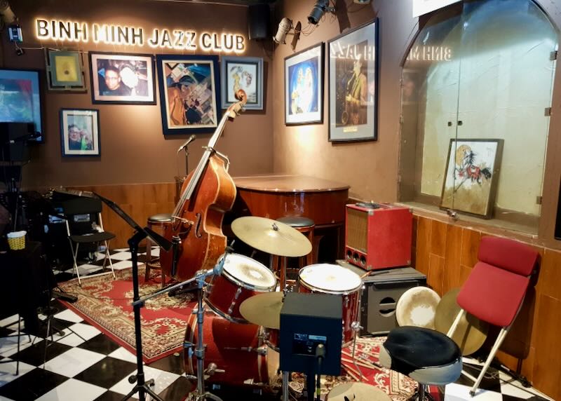 A cello, drum set, amplifier, and piano sit in a corner of a performance club.