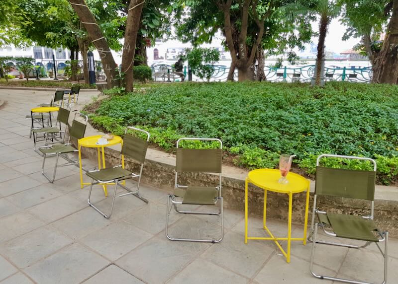 Outdoor chairs and yellow tables sit next to a green patch and the lake.