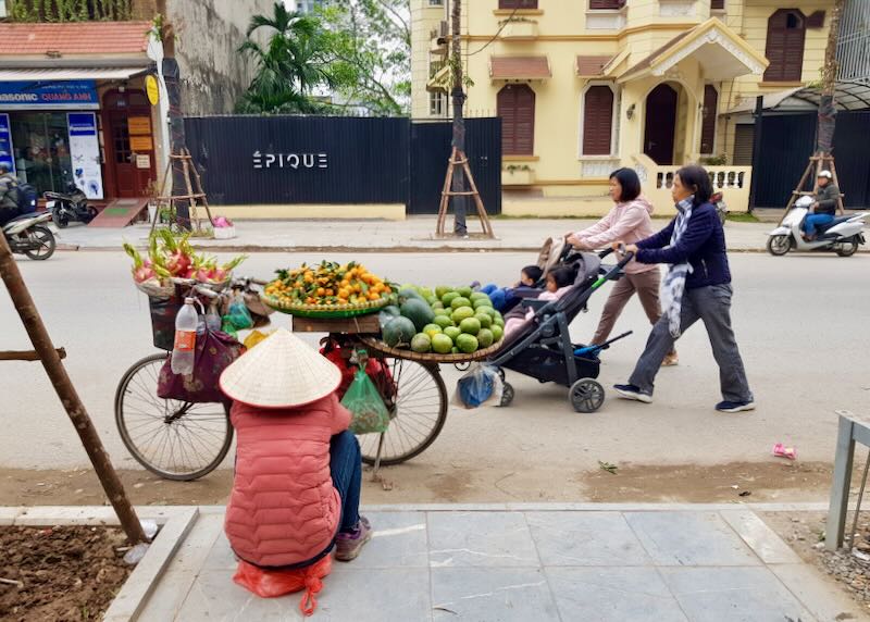 A fruit seller sits by a bicycle loaded with fruit baskets.
