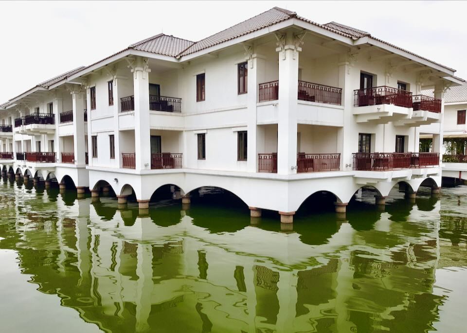 A white hotel with red railings sits above a green lake.
