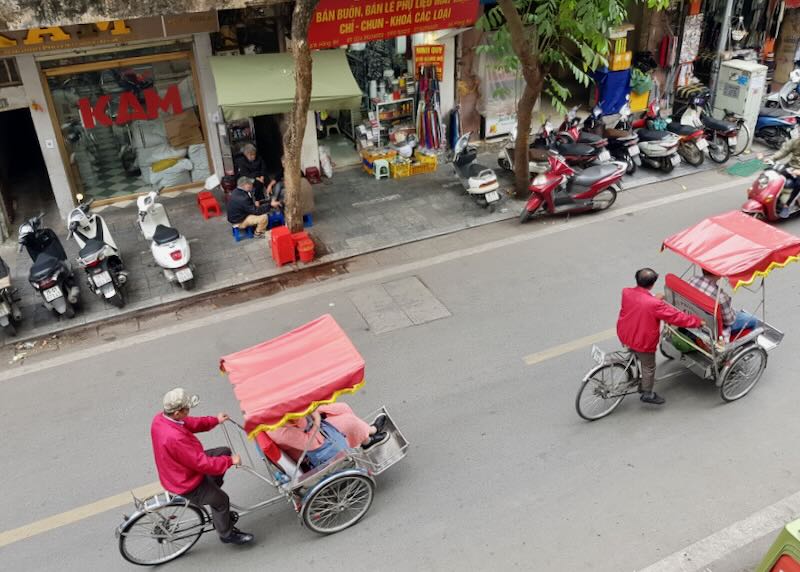 Two men in red jackets pedal rickshaws with customers in a front seat down a street.