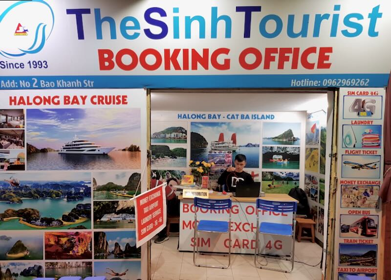 A young man sits at travel desk surrounded by bright blue and red posters advertising day trips.
