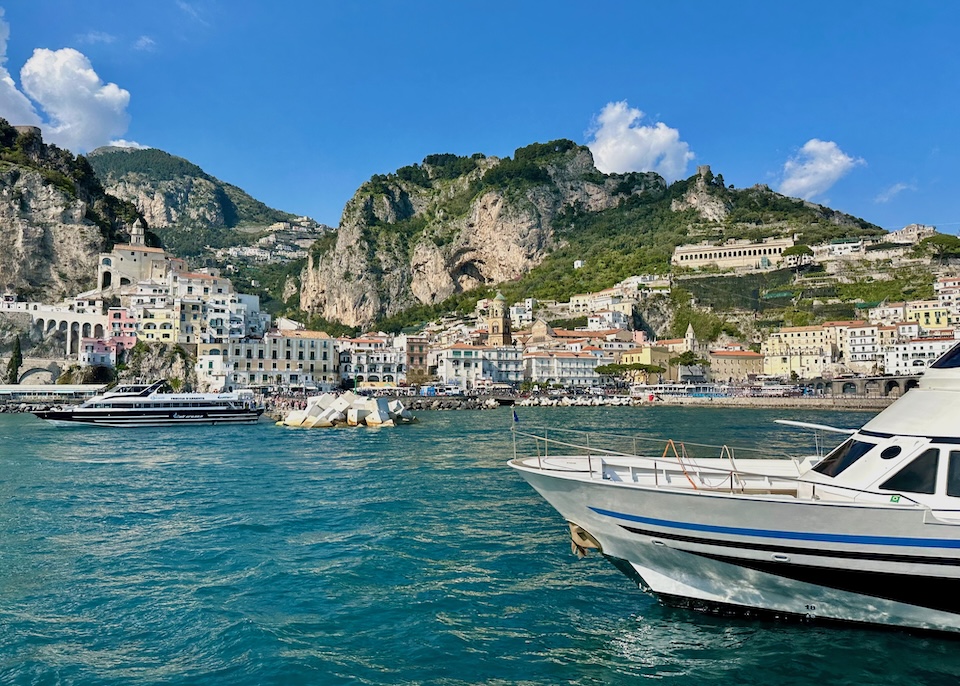 View of Amalfi town from the sea with ferries and boats in front, the cathedral in the center, and mountains rising behind the town