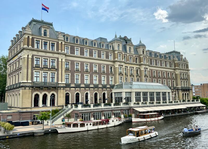 Large ornate hotel on the river Amstel, with boats passing by on a sunny day