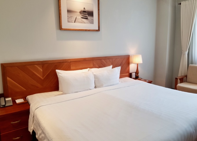 A white linen bed with a orange-stained wood headboard sits in a hotel room.