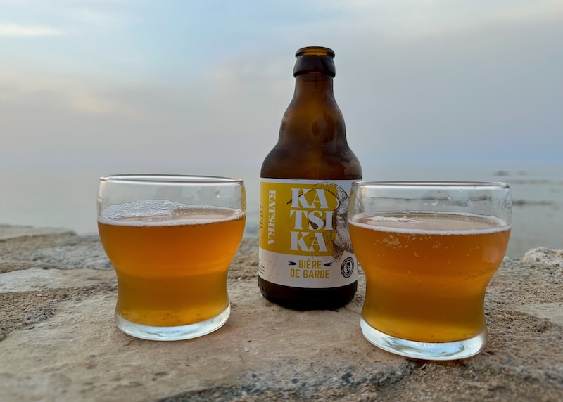 Two glasses of beer and an amber glass beer bottle on top of a stone wall with a stormy sea and cloudy sky behind in Chania, Crete