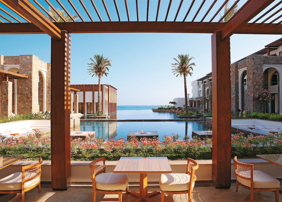 Two central pools on the sea between two stone buildings and in front a dining area and pergola at Amirandes Grecotel resort in Crete