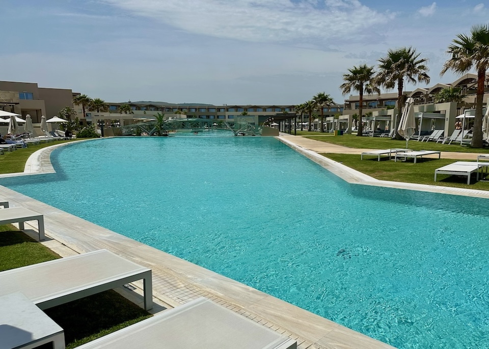 A freeform pool crossed by a bridge set between green lawns at Euphoria resort in Crete