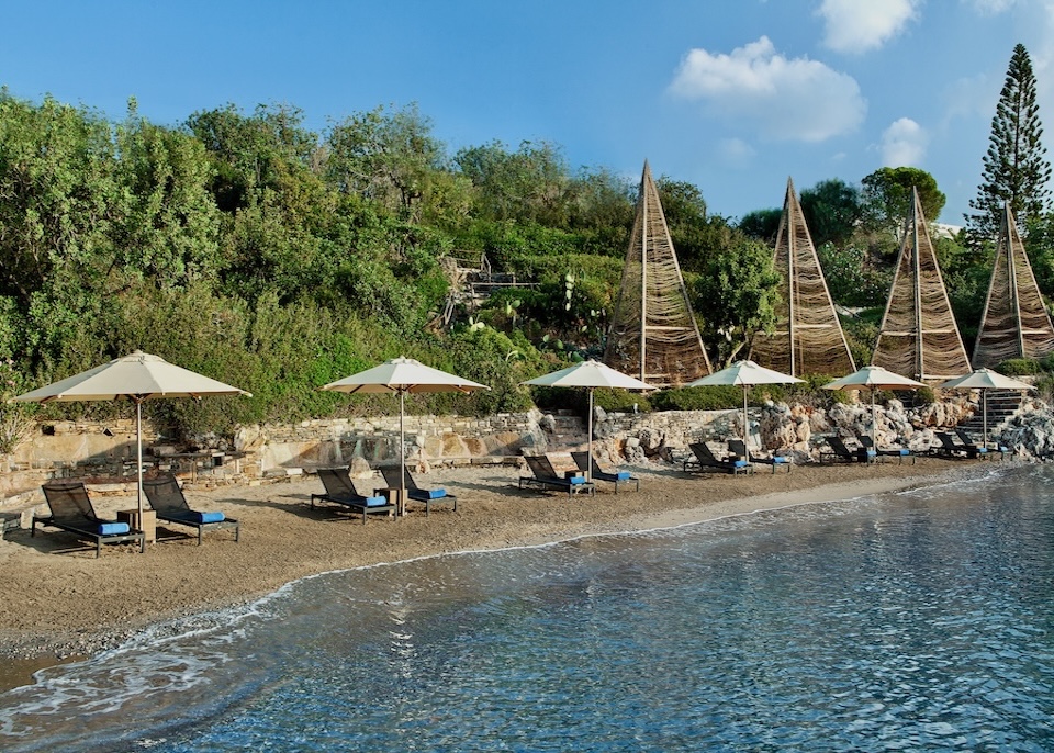 View from the gentle sea toward the beach and forest with sunbeds, umbrellas, and wooden pyramid art at Minos Beach resort in Crete