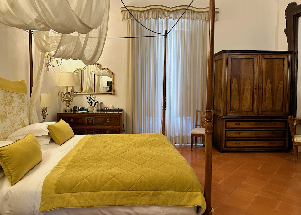 A deluxe room at Palazzo Murat in Positano with a four-poster bed in white and gold with a gauzy canopy and terracotta flooring