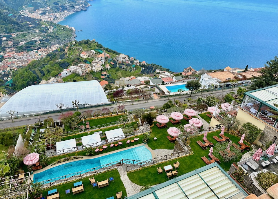 View from the top of Palazzo Avino hotel toward the garden pool, green lawn, and pink-and-white striped umbrellas above the sea in Ravello
